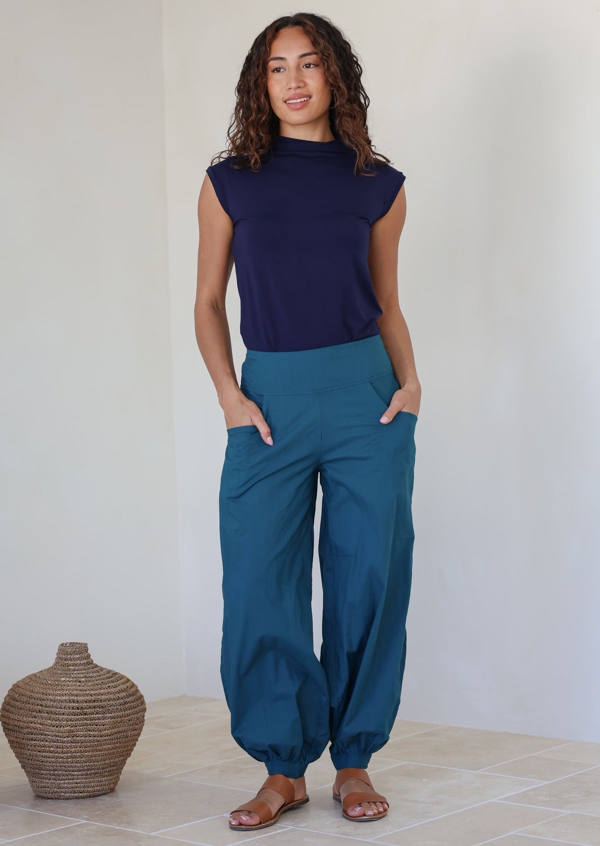 Woman wearing a navy top basic top and pull on style teal pants with cuffed ankles and hands in pockets, standing against a white wall
