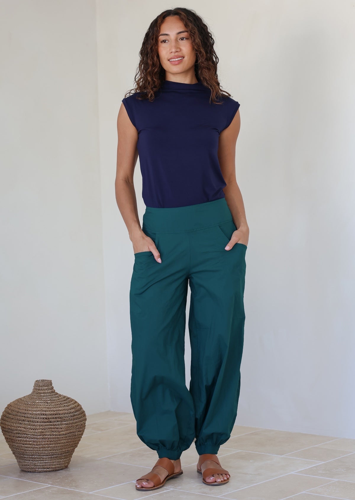 Woman wearing a navy top basic top and pull on style teal pants with cuffed ankles and hands in pockets, standing against a white wall