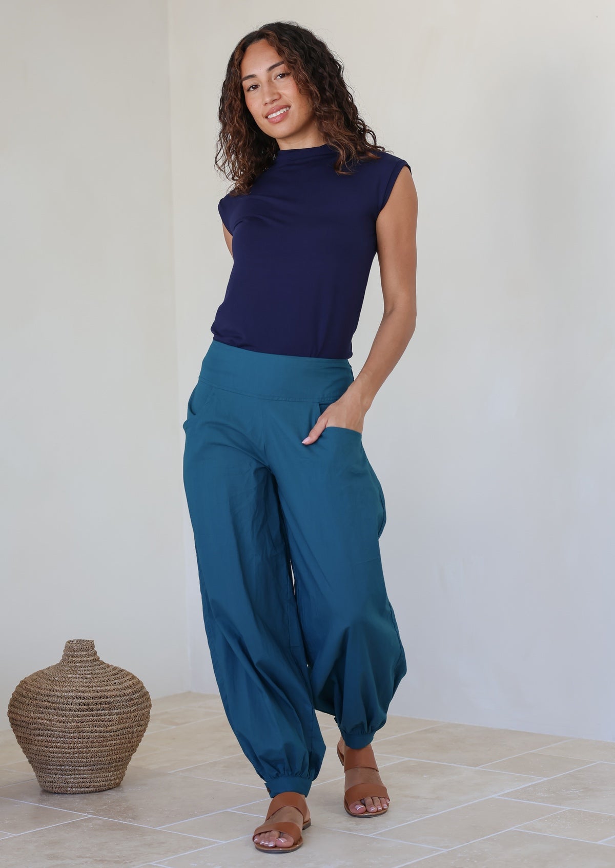 Woman stands with one hand in pocket wearing a genie style pull on pants in deep teal with cuffed ankles, wide leg fit and flat wide waist band that is shirred at the back 