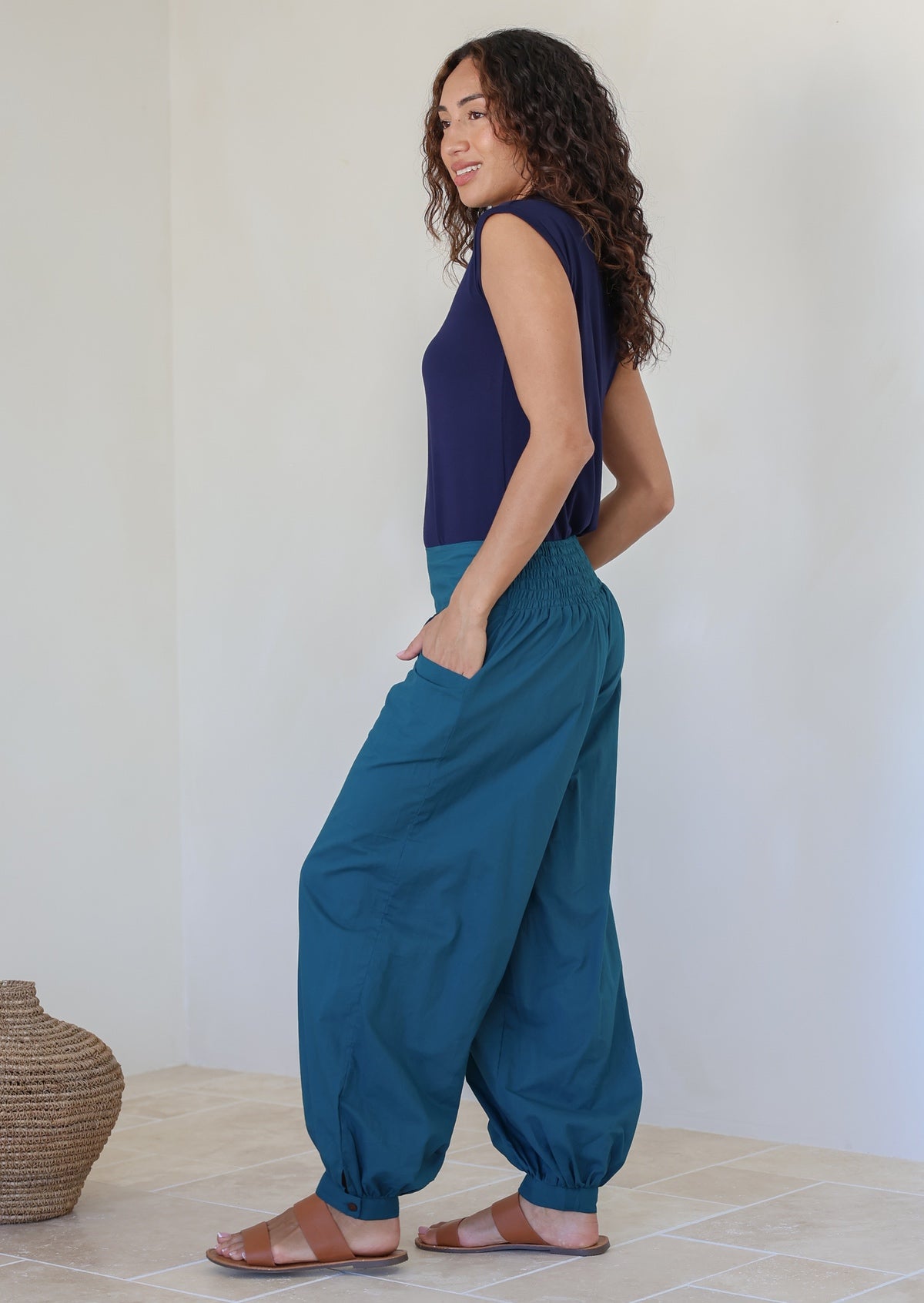 Woman poses with hands in pocket wearing a lightweight deep teal cotton pants with cuffed ankles and elastic at back waist