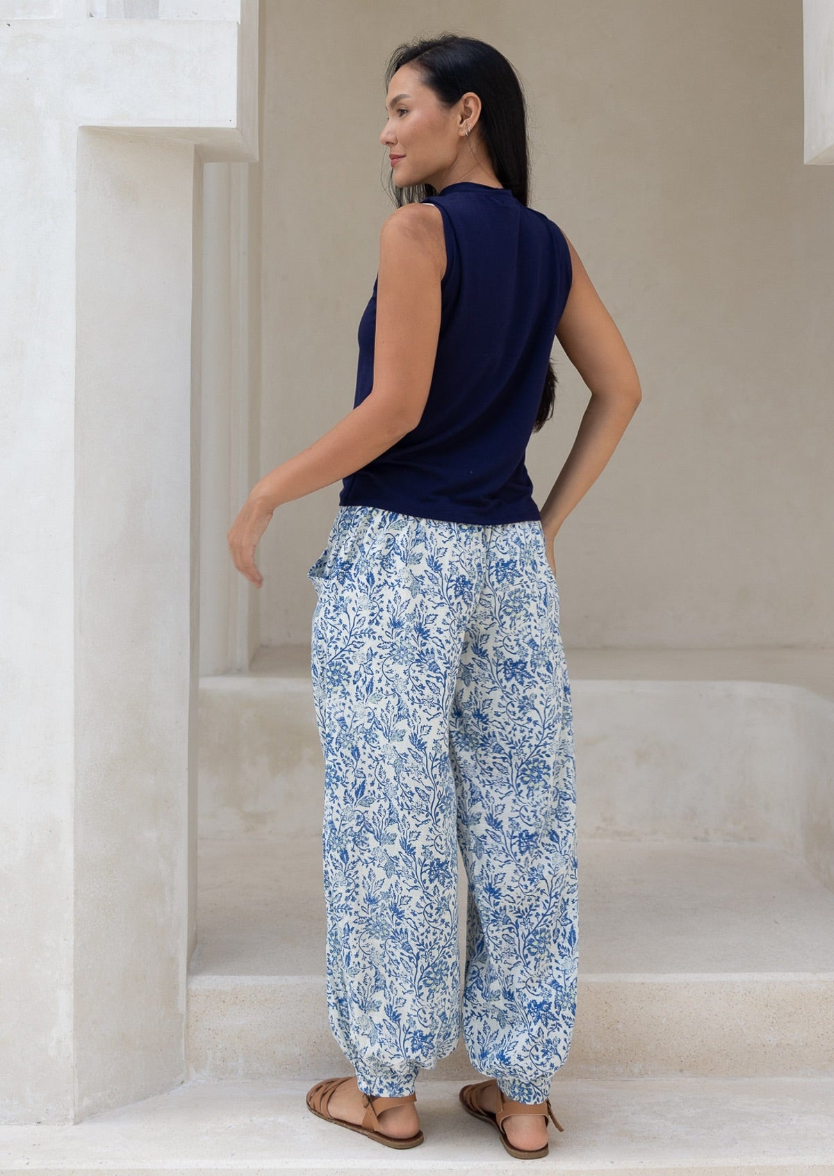 Back view of woman wearing a navy sleeveless top and white based blue floral cotton pants paired with tan sandals