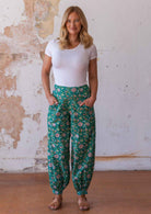 Woman stands in cotton harem pants in floral print on a jade green base with hands in pockets