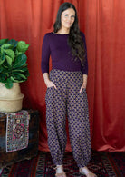 Woman wears cotton buttoned ankle harem pants with floral motif print on a dark purple base 