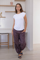 Woman wears cotton floral print with dark purple base harem pants, with buttoned ankle and pockets, paired with a basic white top.