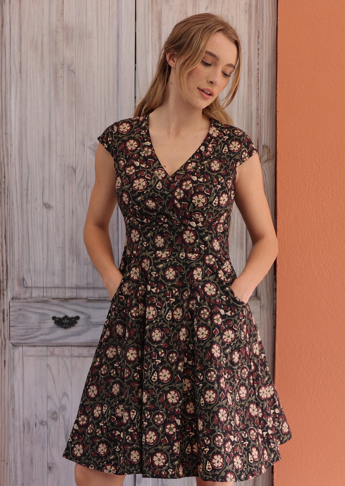 Woman wearing retro inspired cotton knee length dress in maroon and cream floral print on black base, with her hands in pocket.