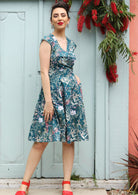 Woman wearing the Alice Dress Verdure, retro cotton sundress with crossover bodice and teal floral print with bright red shoes