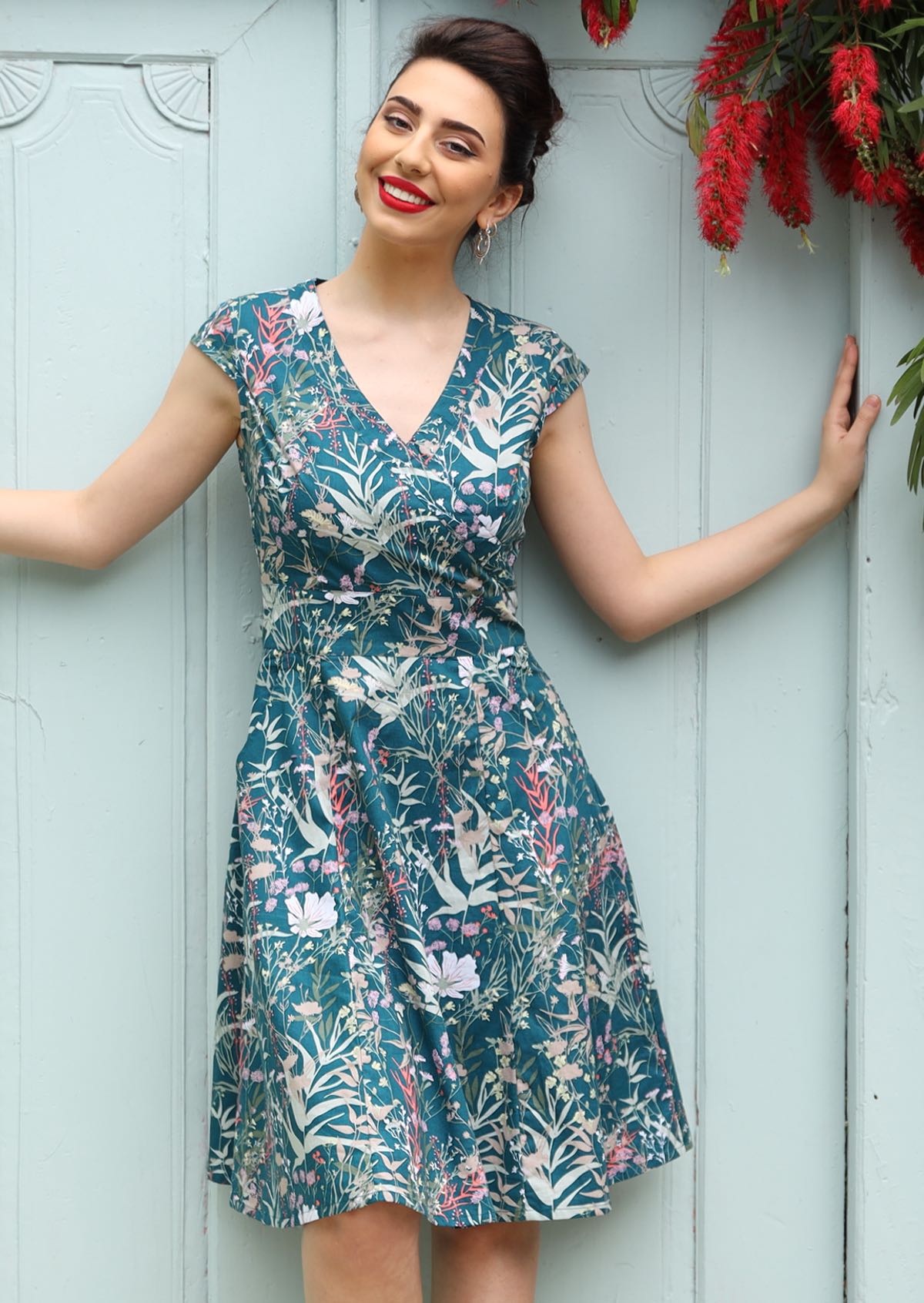 Woman wearing the Alice Dress Verdure, a retro cotton sundress with a crossover bodice, cap sleeves and a teal floral print.