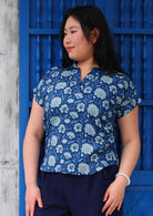 Woman with black hair wearing a blue cotton top with cap sleeves and collar in light blue and white floral print, in front of blue door