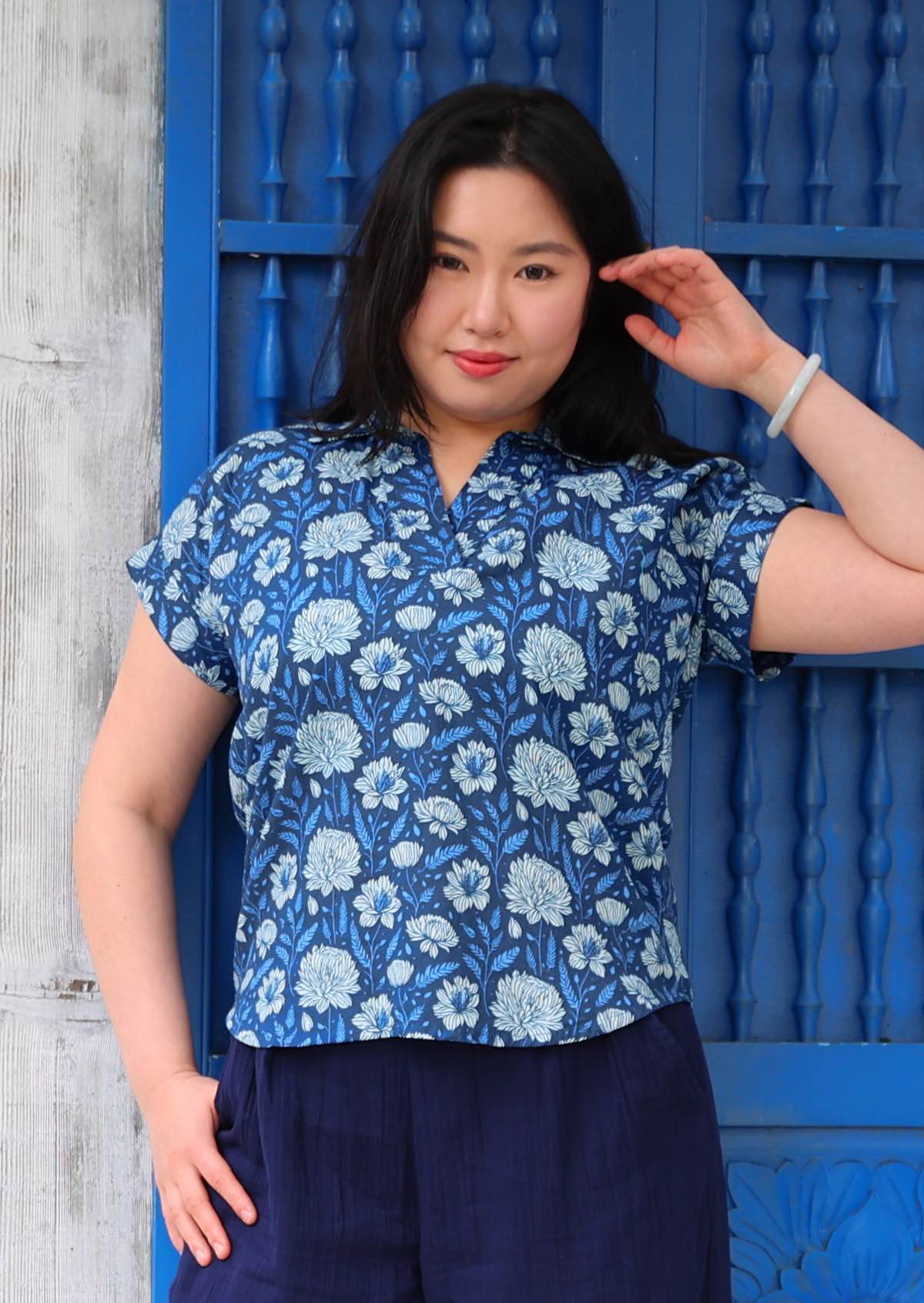 Woman with black hair wearing a blue cotton top with collar and V-neckline in light blue and white floral print, in front of blue door