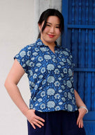 Woman with black hair wearing a blue cotton top with cap sleeves in light blue and white floral print, in front of blue door