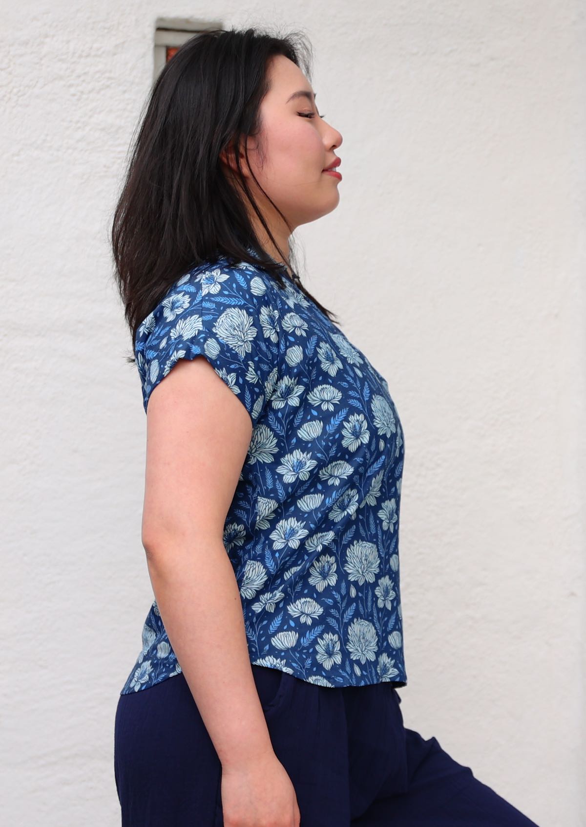 Woman with black hair wearing a blue cotton top with slight curve in bottom hem in light blue and white floral design, in front of beige wall