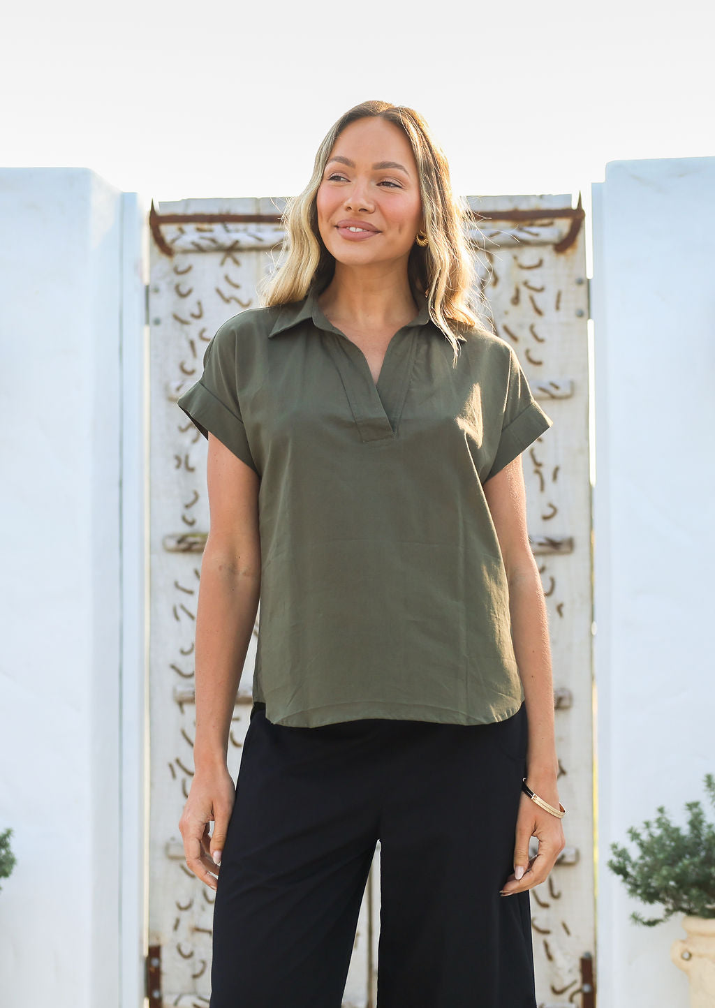 Woman wearing an olive green shirt style relaxed fit top and black pants standing in front of a decorative white wall.