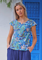 Woman poses in cap sleeved cotton tailored top in multicoloured leaf print on a blue base with hands in pants pockets