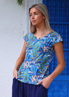 Woman poses to the side in cap sleeved cotton tailored top in multicoloured leaf print on a blue base with hands in pants pockets