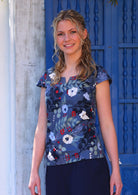 Woman with blonde hair wearing a cotton top with round neckline with keyhole cutout, in blue, white, grey and red floral print, in front of blue door