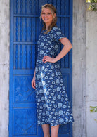 Model standing in front of blue door with hand on hip wearing pale flowers on a navy base 100 percent cotton sundress