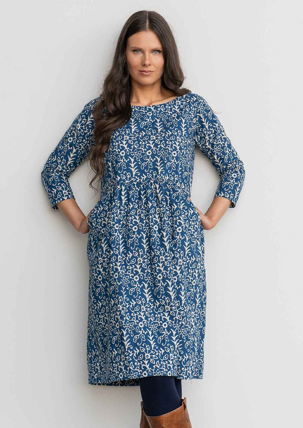 Woman in blue cotton, floral, knee length sundress with sleeves,  with hand in pockets leaning against wall 