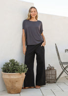 Woman wearing a dark grey stretch rayon short sleeved batwing top and black pants standing against a white wall