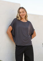 Woman wearing a dark grey soft stretch rayon batwing top and black pants against a light-coloured wall.