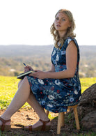 Woman sits on stool in knee length blue floral dress with cap sleeves and cinched waistline