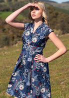 Woman peers into distance wearing blue floral cotton dress with modest V-neckline, cap sleeves, cinched waist with two box pleats