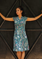 Woman wearing the Billie Dress Verdure, a structured cotton sundress with cap sleeves and a teal wildflower print in front of black door