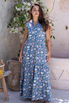 Woman wearing a sleeveless cotton maxi dress with buttoned bodice, 3 tiered maxi skirt, mandarin collar in dusty blue based floral print.