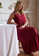 Woman sits on chair wearing cotton sleeveless maxi dress in deep red colour, made from two layers of cotton gauze basted together