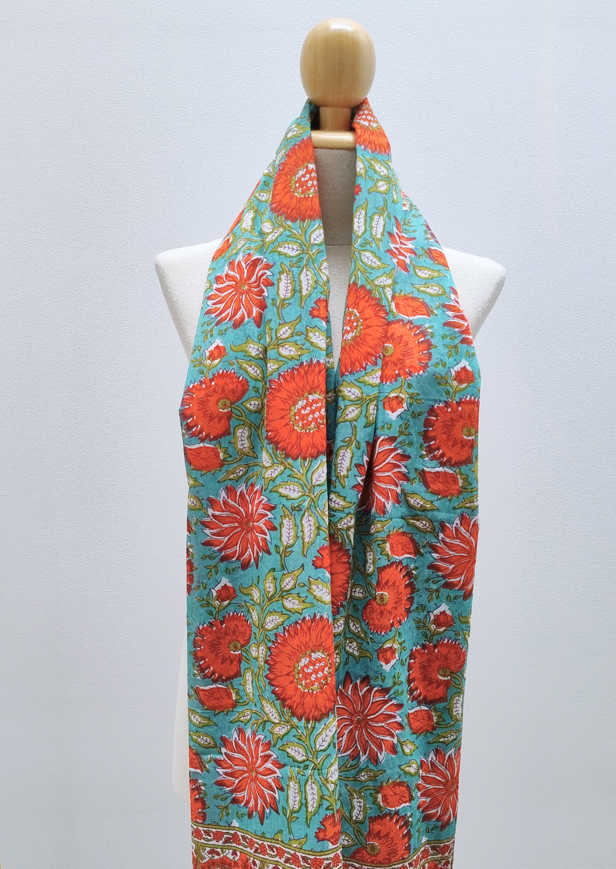 A vibrant scarf with a turquoise blue background and a bold red-orange floral block print, displayed on a white mannequin torso.