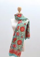 A vibrant scarf with a turquoise blue background and a bold red-orange floral block print, displayed on a white mannequin torso.
