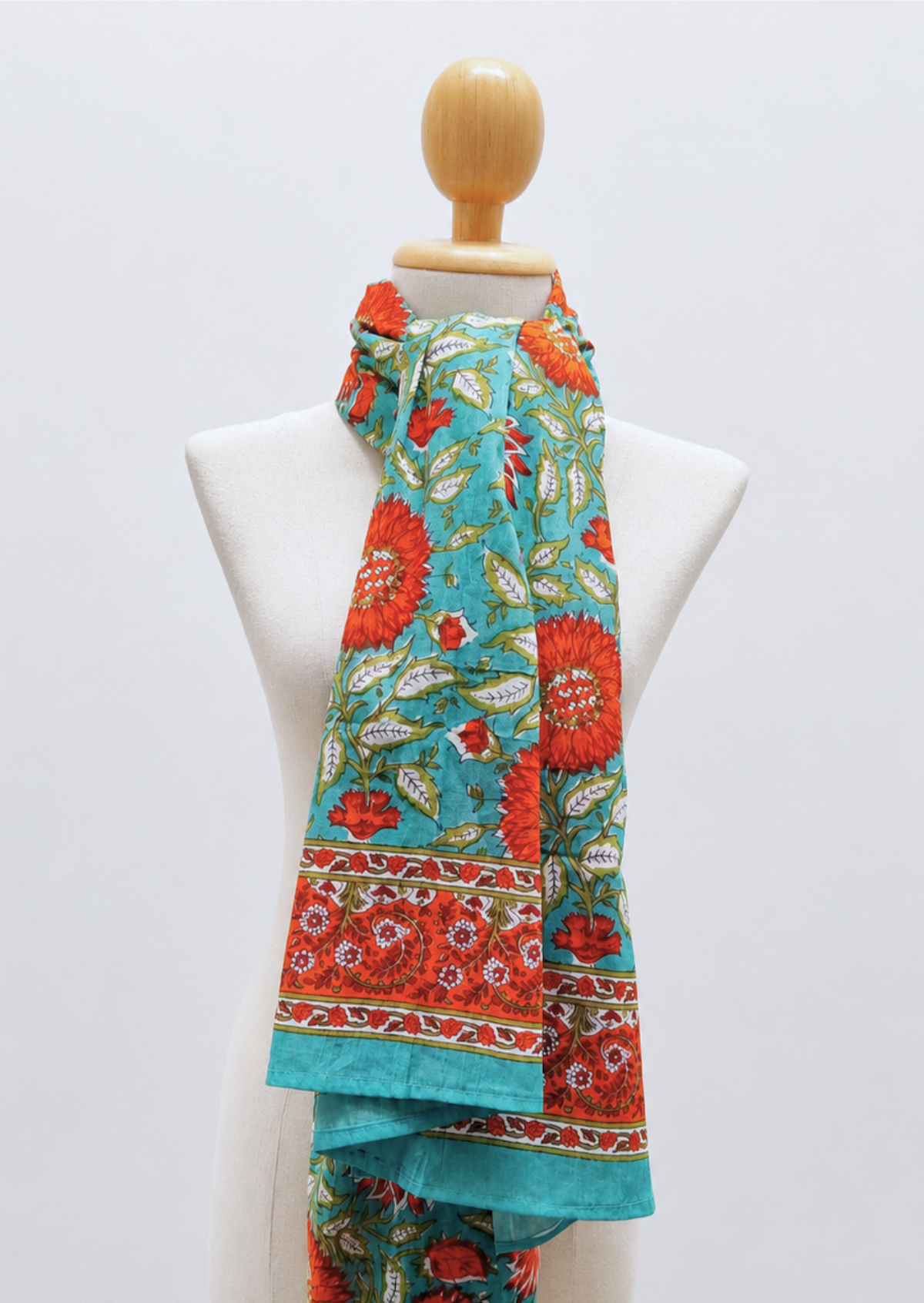 A vibrant scarf with a turquoise blue background and a bold red-orange floral block print, displayed on a white mannequin torso.