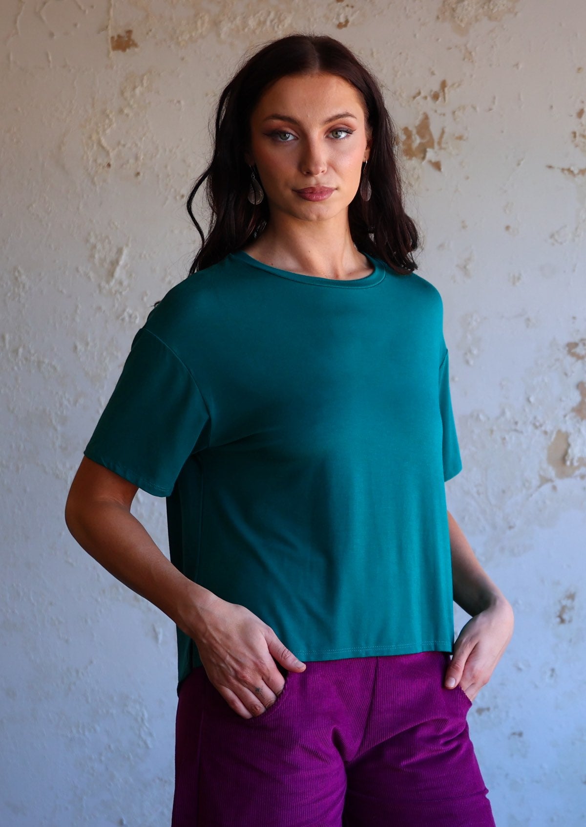 woman wearing loose fit teal blue t-shirt over purple pants with hands in pockets
