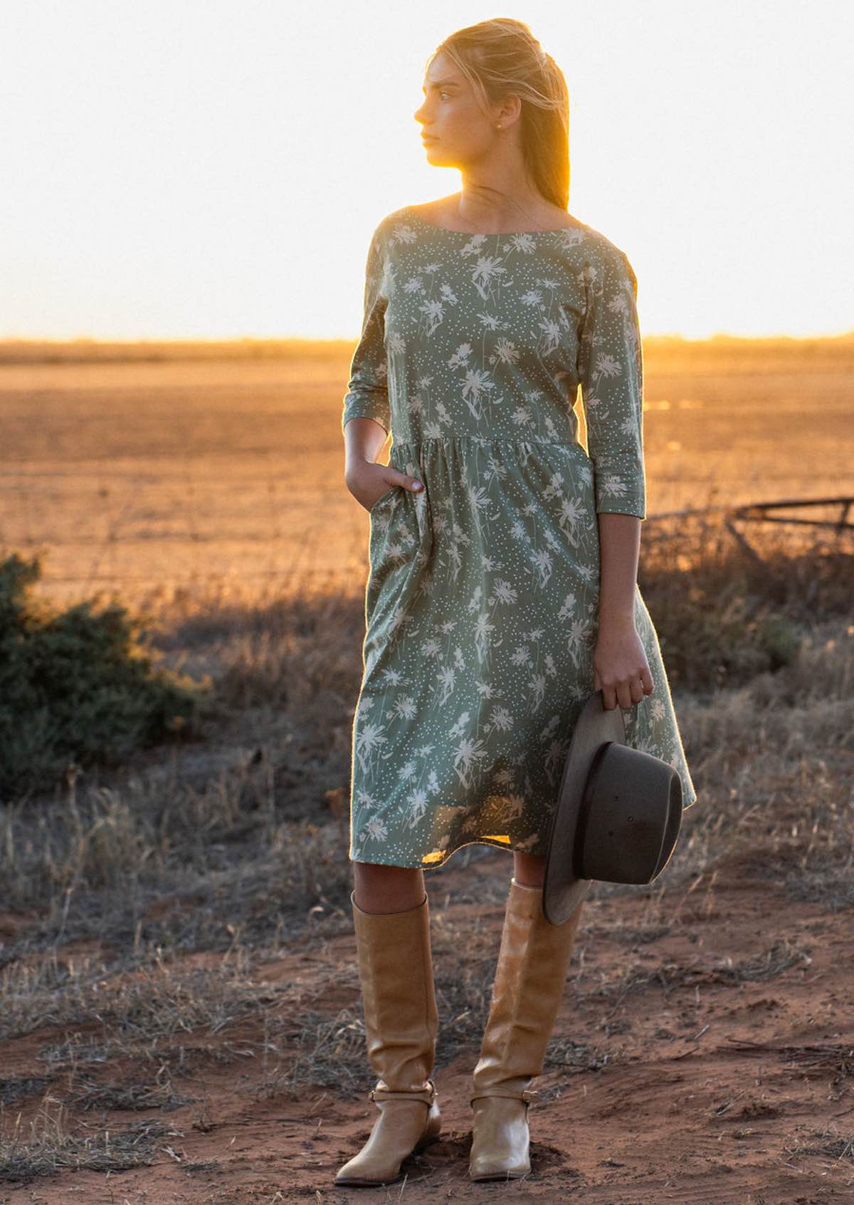 Woman stands in front of setting sun in 100% cotton, over the knee length dress with boat neckline, 3/4 sleeves and pockets