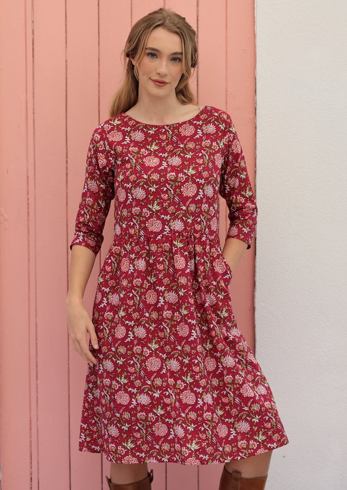 Model standing in front of pink background wears a knee length cotton dress with 3/4 sleeves, round neckline and pockets.
