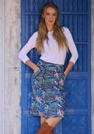Woman wears cotton pencil skirt in leaf print on blue base with contrasting trim around the waistband and pockets, paired with a white top