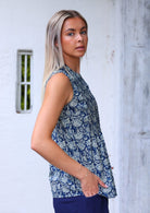 Woman stand sideways in cotton sleeveless blouse in blue floral print, with her hands in pockets of cotton pants