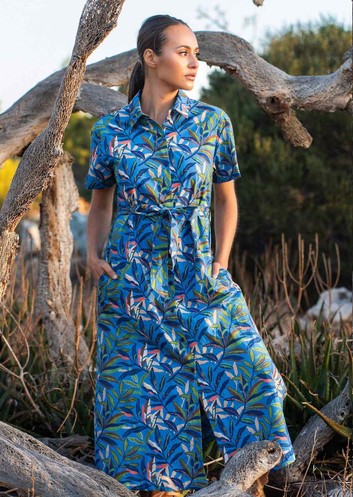 Woman wears cotton shirt dress with collar, in blue based leafy print, with her hands in pockets