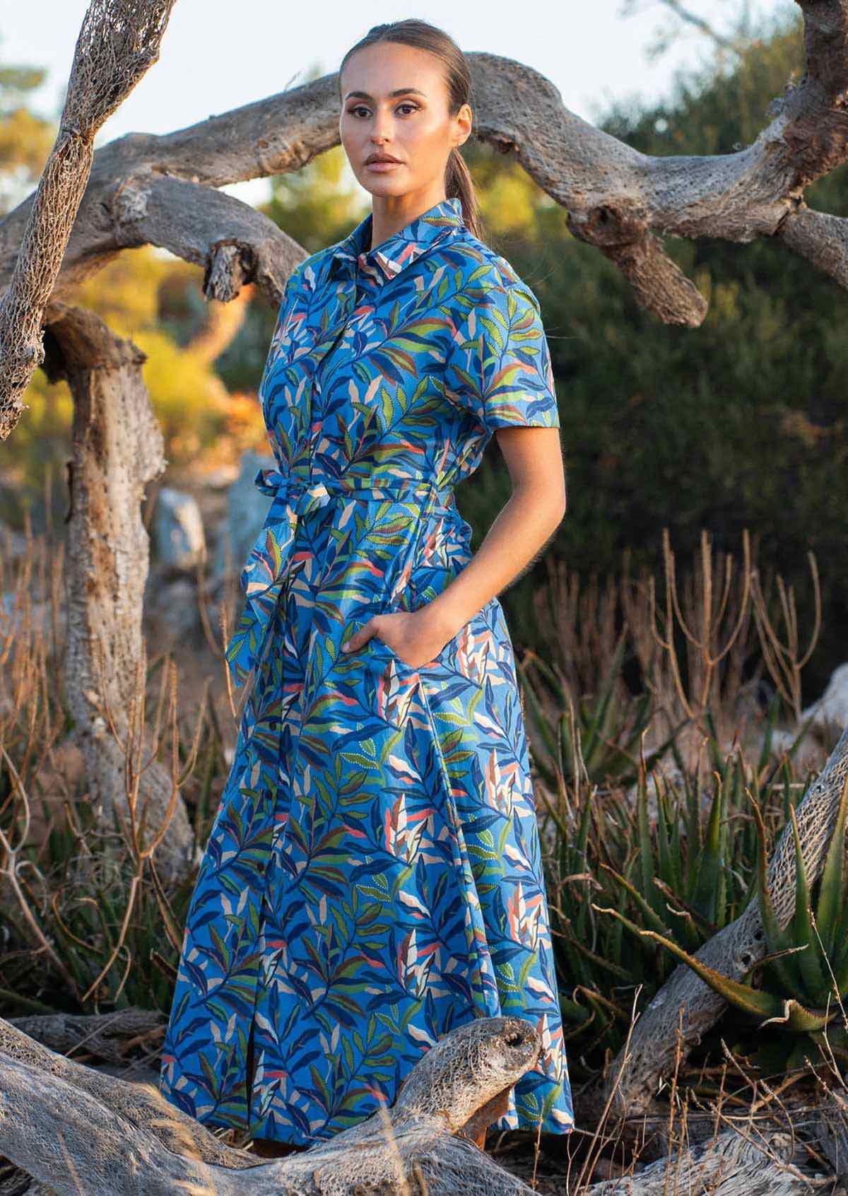 Woman wears cotton midi length short dress in multi coloured leafy print on a blue base with her hands in hidden side pocekts