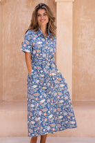 Woman standing in front of beige wall wears shirt style long dress in dusty mid blue with coral pink, lemon yellow and green floral print.