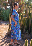 Woman stands sideways in cotton leafy print on blue base dress with hidden side pockets