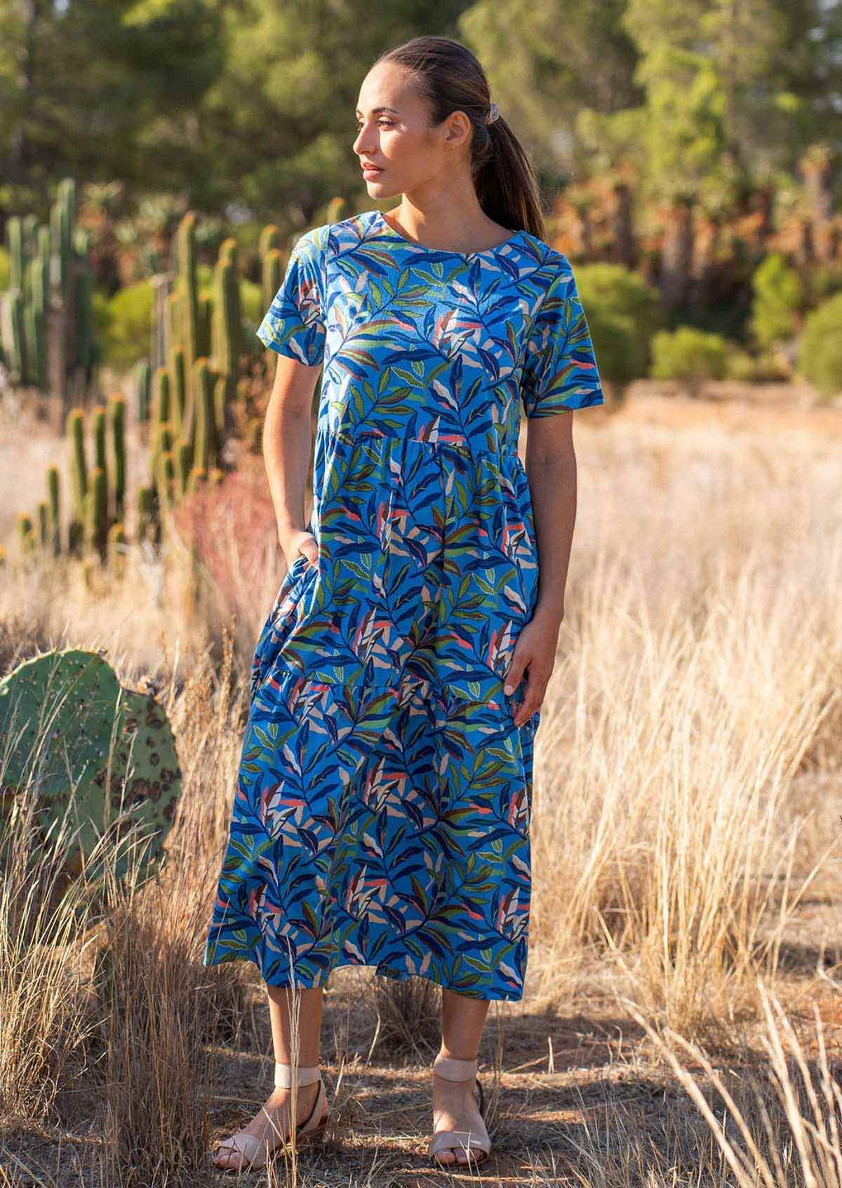 Woman wears cotton midi length tiered dress in multi-coloured leafy print on a blue base