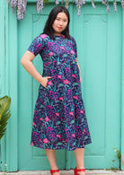 Model with hands in pockets wearing tiered long pink and purple floral on dark blue base 100 percent cotton sundress 