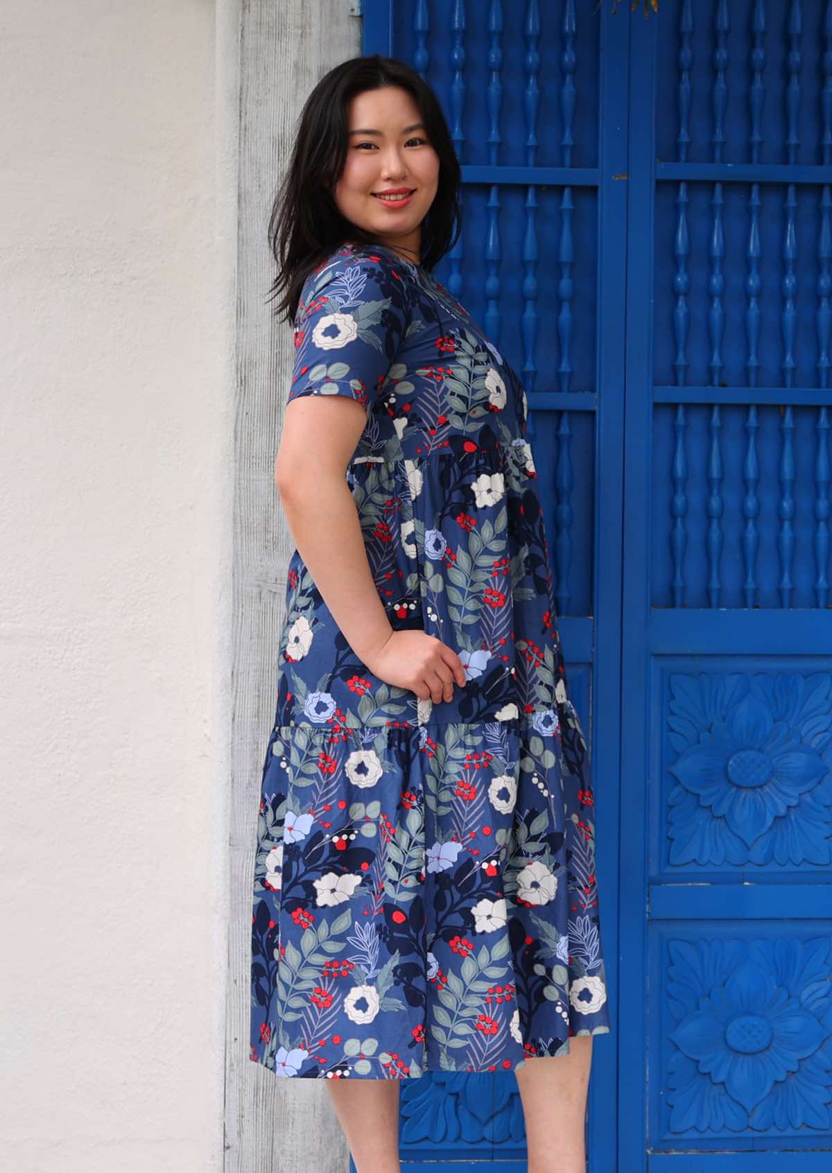 Woman with dark hair wearing a blue cotton dress with white and blue floral designs, hand in pocket in front of blue door