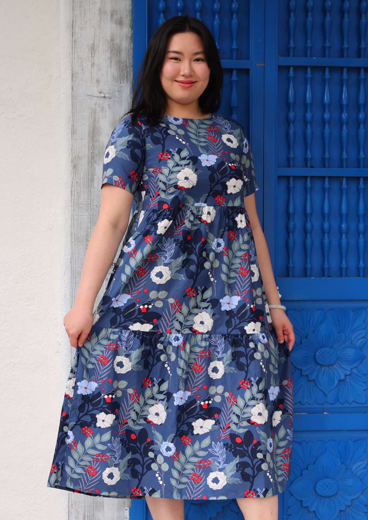 Woman with dark hair wearing a  blue floral cotton dress with white and blue flowers, in front of blue door