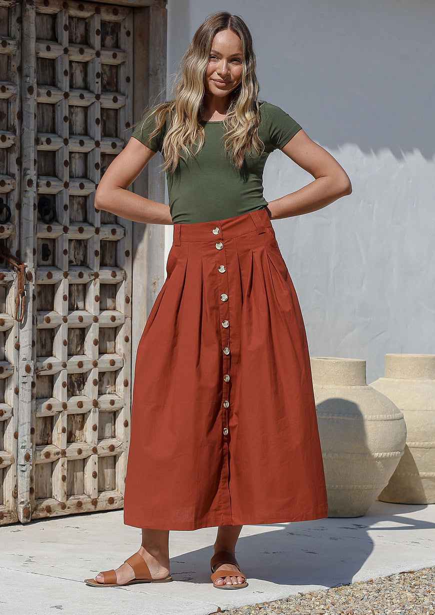 Woman wearing a green basic top and rust colored shin length, A line cotton skirt with box pleats, belt loops and button through front