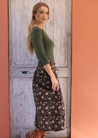 Woman stands sideways in midi length black based cotton skirt with white and maroon floral print, paired with olive stretch rayon top