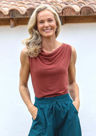 Woman wearing a terracotta coloured sleeveless top with a cowl neckline and teal pants