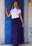 Woman wearing navy blue cotton maxi skirt with white t-shirt in front of bright blue door with tan sandals
