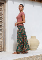 Model standing sideways wears a cotton maxi skirt with wide elasticated waistband in deep green based floral print.