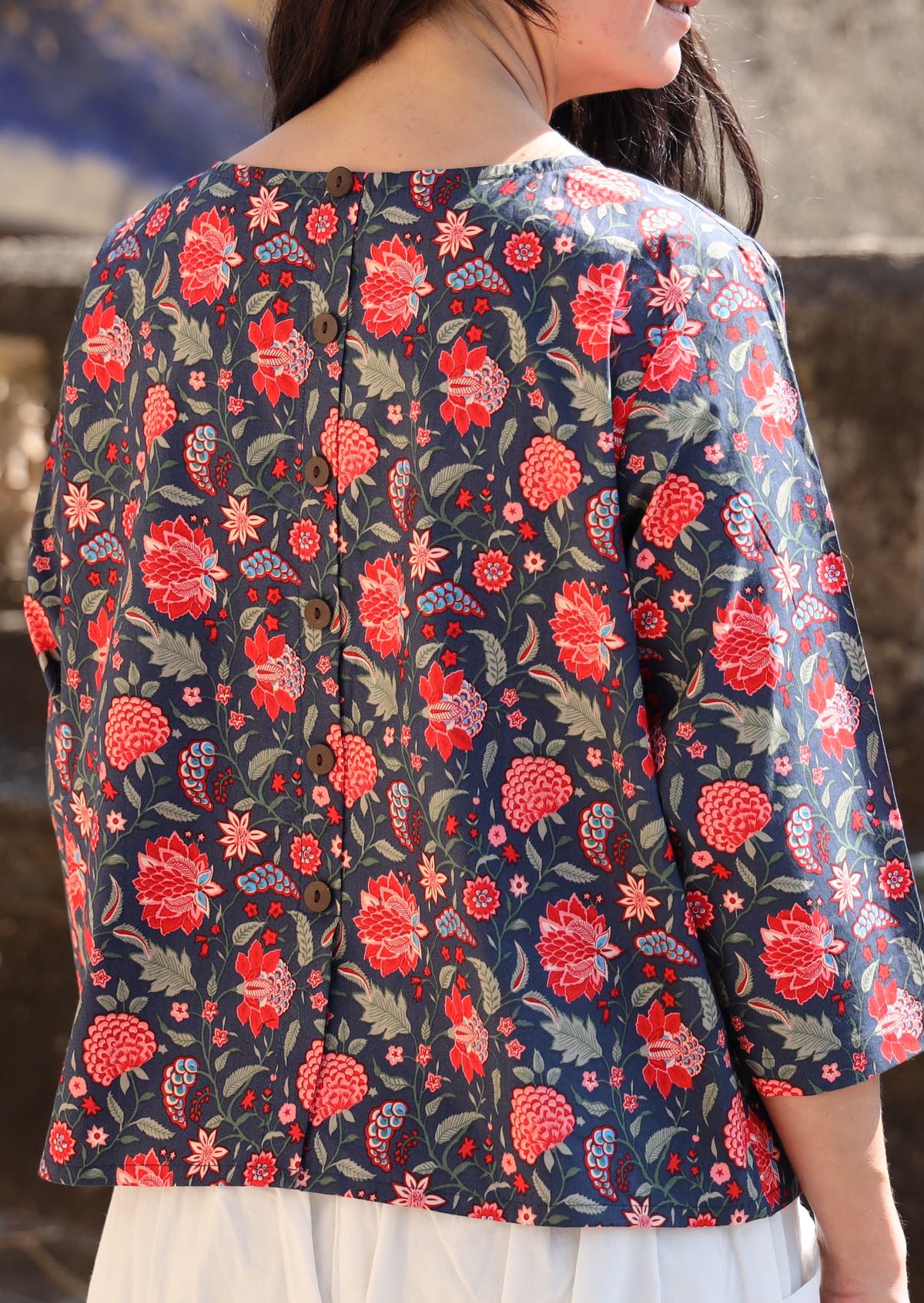 Back of woman in a cotton relaxed fit 3/4 sleeve top with decorative buttons, in a red and green floral print on a blue base 